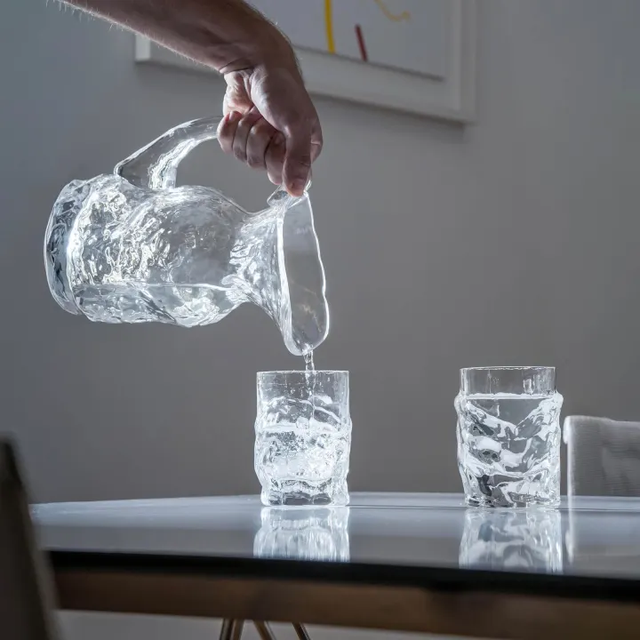 Ruka nalévá vodu z džbánu Liquid Glass do sklenice na skleněném stole